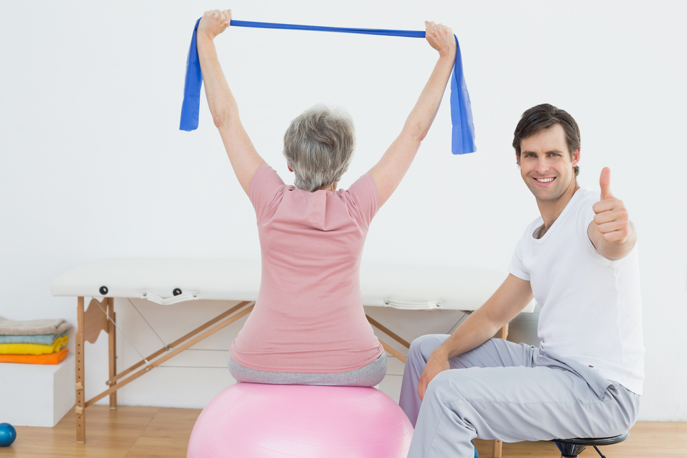 Physical therapist gesturing thumbs up besides senior woman on yoga ball